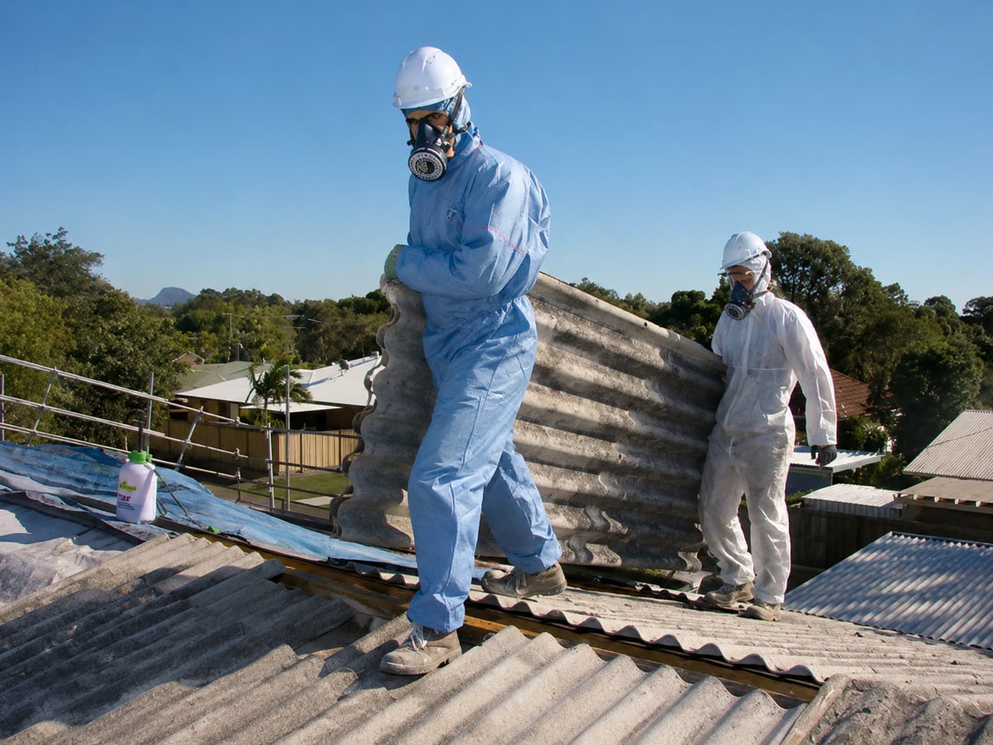 Two operatives in full PPE carefully carrying a corrugated asbestos cement roof sheet across a residential roof in Surrey