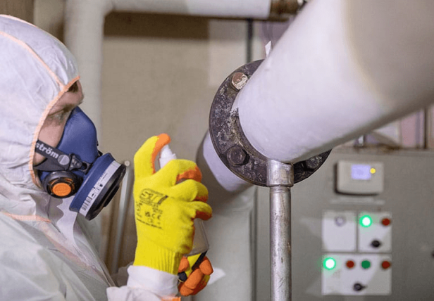 Licensed operative in full PPE inspecting and treating asbestos pipe lagging on a heating system in a Surrey commercial property