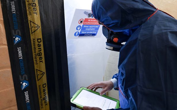 Surrey Asbestos operative in full PPE reviewing documentation at a sealed asbestos hazard enclosure entry point in Surrey